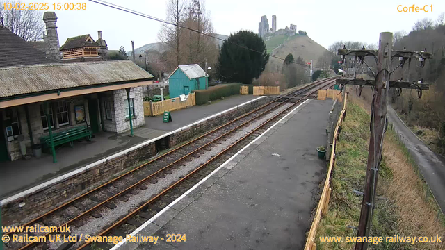 A railway station platform under cloudy skies. In the foreground, the platform is made of stone with a small concrete path, flanked by wooden fencing on one side. There are several rail tracks running through the center, with a light gravel surface in between. On the left, there is a green bench and a stone building with a sloped roof and chimney, possibly serving as a ticket office or waiting area. In the background, to the right, a green shed and a sign indicating "Way Out" are visible. Rising up behind the station, a hill features a castle ruin, partially obscured by trees.