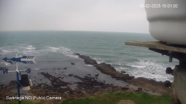 A view of the coastline on a cloudy day, with rough waves crashing against rocky shores. In the foreground, a weather vane indicates the direction of the wind, and there are partially visible rocks in the water near the shore. The sea appears grayish with foam from the waves, and the sky is overcast. There is a timestamp in the upper right corner indicating the date and time of the image.