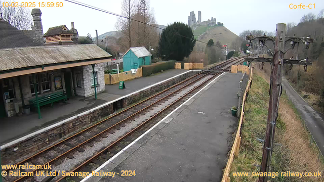 A view of a railway station platform with tracks running along the bottom. A stone building with a sloped roof is on the left, featuring benches and a sign. In the background, there is a small green shed and a line of trees. A hill with castle ruins can be seen in the distance against a cloudy sky. The platform is bordered by a wooden fence, and there are power lines and a pole on the right side of the image.