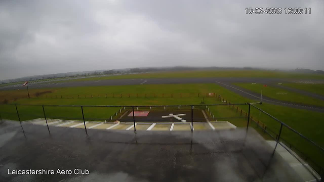 A cloudy sky above a grassy airfield viewed from a raised platform. The foreground features a white structure resembling a cross on the ground, next to a red square shape. In the background, there are several runways and a faint outline of trees or buildings in the distance. The overall scene appears overcast with low visibility.