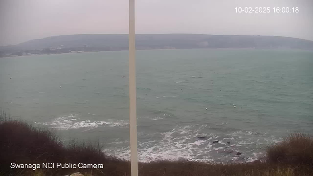 A coastal view over water on a cloudy day. The water appears choppy and is colored greenish-blue, with gentle waves lapping against rocky areas. In the distance, there are faint outlines of hills and a few buildings along the shore. The sky is overcast, and the scene conveys a calm but cool ambiance. A white vertical pole is present in the foreground, and the image includes a timestamp in the upper right corner indicating the date and time.