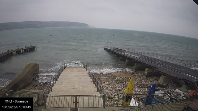 A view of a coastal area with grey skies and choppy waters. In the foreground, there are steps leading down to the water, flanked by a stone and metal railing. To the left, a wooden pier extends into the ocean, while to the right, a metal ramp leads to another platform over the water. Near the bottom of the image, there are several colorful kayaks (yellow, blue, and red) stored on a rocky shore. In the background, distant cliffs can be seen fading into the horizon.