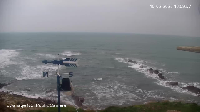 A view of the ocean under a cloudy sky, with waves crashing against rocky formations in the water. In the foreground, there is a weather vane displaying directional indicators, with the letter "N" for north and "S" for south prominently visible. The coastline is partially visible, with rocky outcrops along the water's edge. The date and time are displayed in the upper right corner.