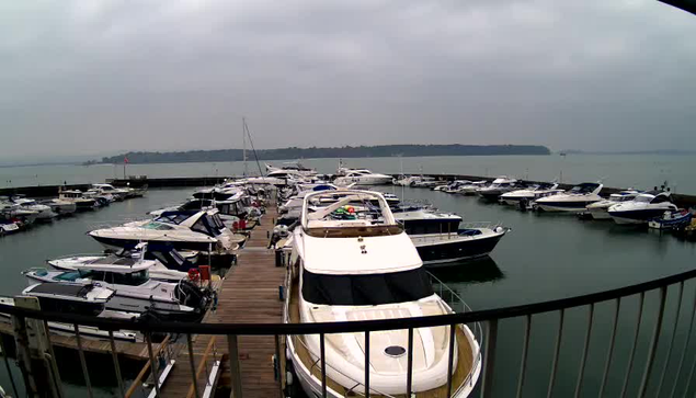 A harbor filled with several motorboats and yachts, anchored neatly in a circular formation with a wooden dock in the foreground. The sky is overcast, creating a gloomy atmosphere, and the water is calm, reflecting the boats. In the background, a shoreline with trees is visible, but the details are muted by the cloudy sky.