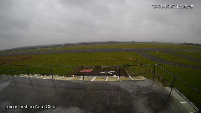 A view from a webcam positioned at an airfield. The scene is overcast with gray clouds. In the foreground, there is a railing and a wet surface, suggesting recent rain. Below, a helipad is marked with white paint in the shape of a cross, surrounded by a red square. In the background, a large expanse of green grass is visible, along with a runway marked by asphalt. There are several light poles and a few distant trees on the horizon, indicating a rural setting.