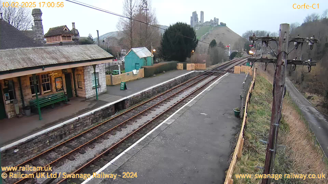 A view of a train station with a stone platform and a roofed waiting area. To the left, there's a long green bench under the shelter. In the background, a small blue building is visible, along with a wooden fence and signs indicating the way out. The scene is set against a hillside with ruins atop, partially obscured by trees. The track features multiple rails and gravel, extending toward the right. A wooden utility pole with wires stands prominently on the right side. The sky is overcast, suggesting a cloudy day.