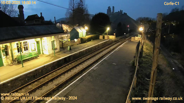 A dimly lit railway station at dusk. The platform is mostly empty, featuring a green bench and a sign that says "Way Out." On the left, there's a stone building with a sloped roof and windows illuminated by warm light. In the background, there are distant hills, and at the top of the hills, the silhouette of a castle is visible against the twilight sky. The tracks run parallel along the platform, leading into the distance. Street lamps cast a gentle glow along the station.