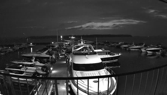 A nighttime scene of a marina with various boats docked. The image is in black and white, showing multiple yachts and smaller boats moored along wooden walkways. The water is calm with some gentle ripples reflecting faint light from distant sources. The sky appears dark with scattered clouds, and no clear moonlight is visible.