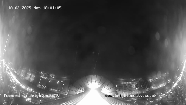 A black and white image from a webcam captures a view at dusk. The foreground features a bright, illuminated area at the center, which appears to be a building or structure with an arch shape. On either side, there are faint outlines of lights and buildings, suggesting a bustling city environment. The top of the image displays the date and time: February 10, 2025, at 18:01:05. The bottom left corner has text indicating it is powered by Brighton CCTV. The overall scene conveys a sense of urban activity and illumination.