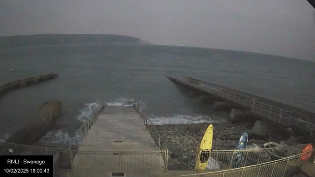 A wide view of the shoreline at Swanage during dusk. The scene features choppy waves in the ocean, with the horizon blending into a cloudy sky. A wooden jetty extends into the water from a concrete path lined with railings. On the right side, two kayaks, one yellow and one blue, are positioned on the ground near the water's edge. The rocky shoreline is visible beside the jetty.