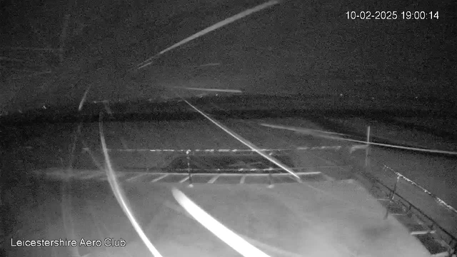 A dark, nighttime view of an airport tarmac with faint streaks of light across the image, suggesting movement. The ground is mostly empty except for a few faint lines indicating the layout of the tarmac and parking area. In the background, a low horizon can be seen against the dark sky. The bottom left corner displays the text "Leicestershire Aero Club" in white. A timestamp indicates the date and time.