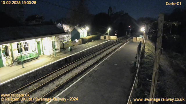 A dimly lit train station platform at dusk. To the left, there is a stone building with green trim, featuring large windows and a bench in front. A sign on the wall indicates services. There are wooden fences and a pathway leading to the left, where a small building is visible. The platform has two sets of railway tracks running parallel, and several lights illuminate the area. A sign indicating “Way Out” is also present on the platform. In the background, trees are silhouetted against the twilight sky.