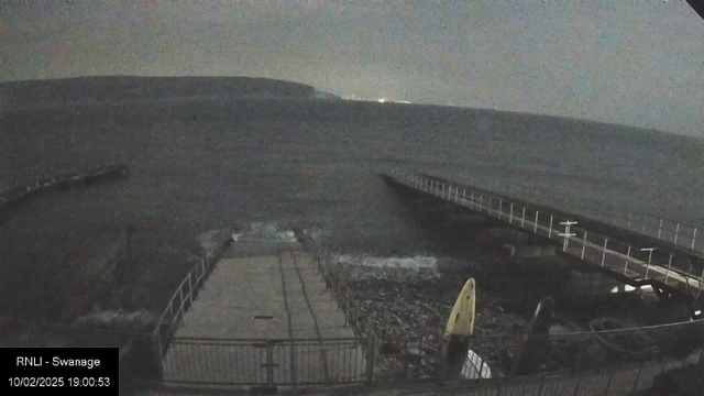 A dark, overcast scene depicting a coastal area. In the foreground, a wide, concrete pier extends into the water, surrounded by a metal railing. Waves gently lap against the edge of the pier. To the left, a portion of the pier curves away from view. The water appears choppy, reflecting faint light from distant sources, while a faint outline of hills is visible in the background. A surfboard, yellow in color, leans against the pier on the right side, suggesting a coastal activity. Overall, the scene conveys a calm yet slightly moody atmosphere.