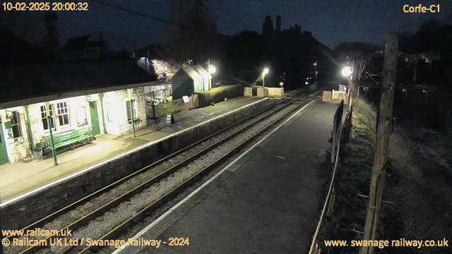 A dimly lit railway station platform at night. The platform features a stone building with large windows and a green bench. There is a sign saying "WAY OUT" in front of a wooden fence. The tracks are visible with a few lights illuminating the area. In the distance, there is a hill with structures silhouetted against the night sky.