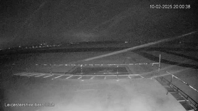 A dark, nighttime scene captured from a webcam at Leicestershire Aero Club. The foreground features a parking area with faint white lines marking spaces, bordered by a low fence. In the background, there are distant lights, suggesting the presence of buildings or other structures. The overall atmosphere is quiet and somewhat empty, with a clear sky overhead.