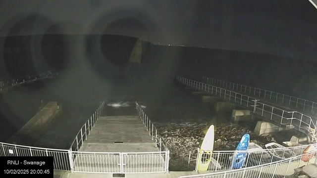 A nighttime view of a coastal area featuring a wooden walkway leading down to the water. The scene includes two kayaks, one yellow and one blue, positioned on the shore near a rocky area. The water is visible at the bottom of the image, with gentle waves breaking. In the background, a dark silhouette of land is outlined against the sky. The image is taken from a webcam, with a timestamp in the bottom left corner indicating it is 20:00:45 on October 2, 2025.