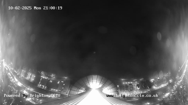 A black-and-white webcam image showing a nighttime scene. The view captures a street with bright lights indicating vehicles, possibly a busy urban area. A prominent structure is visible in the center, with a rounded roof and illuminated edges. The date and time are displayed at the top left corner of the image. At the bottom left, the text "Powered By Brighton CCTV" is visible, and the website URL is displayed at the bottom right.