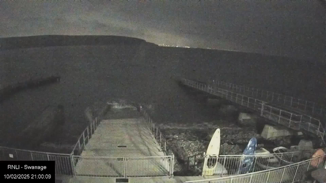 A dark waterfront scene captured by a webcam. A wooden pier extends into the water from the left side of the image. In the foreground, a staircase leads down to the water, surrounded by a fence. Two kayaks, one yellow and one blue, are visible on the right, resting on the ground. The water appears calm, and a distant shoreline is faintly visible in the background. It is nighttime, with an overcast sky and limited visibility. The time and date displayed at the bottom indicate 21:00 on February 10, 2025.