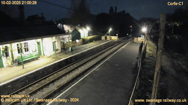 A dimly lit railway station at night. On the left, there is a stone building with several windows and a green bench in front. A sign indicates the way out. The platform is empty, with two sets of railway tracks running parallel, and a wooden fence beyond the platform area. Small lights illuminate the scene, casting shadows, while the outline of a hill and trees can be seen in the background.