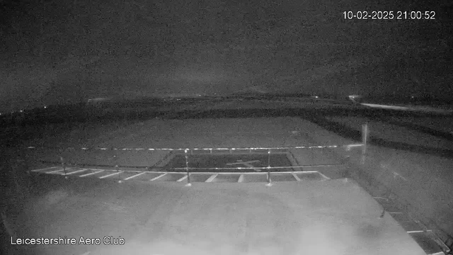 A nighttime webcam image showing a dark, open area with a low fence in the foreground. In the center, there is a helipad marked by a symbol. The background features a view of the horizon, which is faintly illuminated by distant lights, suggesting a rural or semi-urban landscape. The overall scene is primarily dark with minimal contrast. There is a timestamp in the upper right corner indicating the date and time of the image.