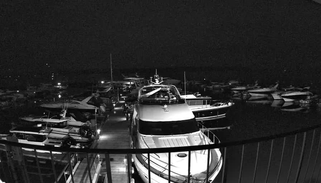 A dimly lit marina at night, filled with various boats and yachts. The scene features multiple vessels docked closely together along a wooden pier. One large white yacht is prominent in the foreground, with its bow facing the camera. Soft lights from the boats and surrounding areas create a subtle glow, contrasting with the dark water and night sky. The atmosphere is tranquil and calm, typical of a nighttime harbor setting.