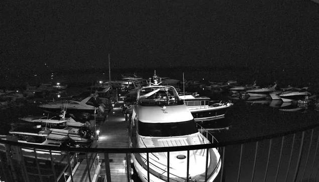 A dark scene of a marina at night, featuring several boats docked in the water. A large white yacht is prominently positioned in the foreground, with other smaller vessels visible in the background. The atmosphere is quiet, with faint lights reflecting off the water and illuminating parts of the boats. The overall setting conveys a calm and serene waterfront environment.