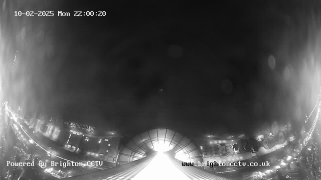 A nighttime webcam view shows a bright, illuminated area in the center, possibly a street or a large building, surrounded by darker surroundings. The top of the image includes a timestamp indicating the date and time, while the lower corners display website and CCTV information. Light reflections and spots appear on the surface, suggesting moisture or glare on the camera lens. The overall tone is black and white, creating a stark contrast between the light and dark areas.