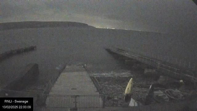 A dark and cloudy seaside scene captured from a webcam. In the foreground, there is a partially visible boat ramp made of concrete with some rocky areas around it. To the right, a wooden jetty extends out over the water, which appears still and quiet. The horizon shows a faint outline of a landmass, and at a distance, a few lights can be seen along the coastline. The overall atmosphere is dim and moody, indicating evening or nighttime conditions.