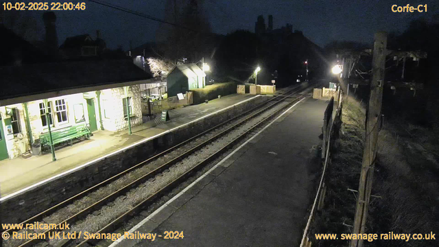 A dimly lit railway station scene at night, featuring a stone station building on the left, with windows reflecting faint light. There are green benches on the platform and signs indicating "WAY OUT." The railway tracks stretch into the distance, with wooden fencing along the edge of the platform. Sparse lighting creates shadows, and the area appears quiet, suggesting it is after hours. In the background, silhouettes of buildings and trees are visible against the dark sky.