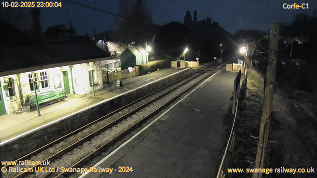 A dark scene shows a quiet railway station at night. The platform is illuminated by various lights. On the left, there is a stone building with green trim, featuring large windows and a green bench in front. A sign indicates the exit path. The tracks are visible, running straight into the distance. To the right, there is a small wooden fence enclosing part of the platform, and a post with overhead wires stands nearby. The background features dark silhouettes of trees and possibly hills, suggesting a rural setting.