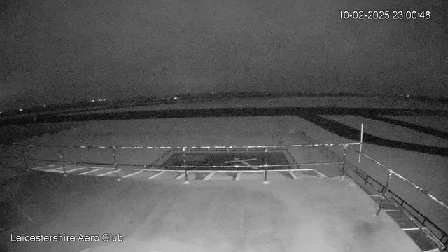 A dark, nighttime scene captured by a webcam, showing a grassy area with a few white lines on the ground, possibly indicating a helipad. A low railing is visible in the foreground, and faint lights are seen in the background, suggesting there are distant structures or trees. The atmosphere is dim, giving a quiet and isolated impression.