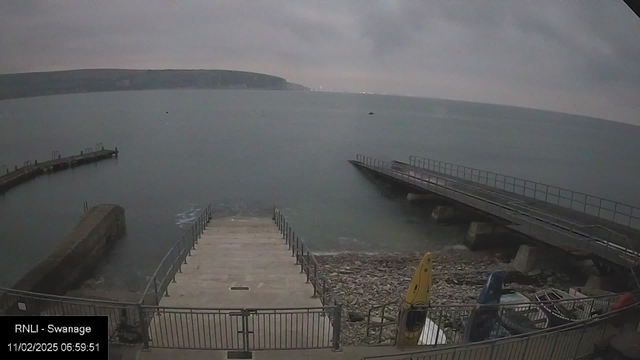 An overcast view of a coastal area at dawn. The foreground features a set of concrete steps leading down to a rocky shore, bordered by a low metal railing. To the left, a curved pier extends into the water, while a straight wooden dock is visible to the right. Several boats are parked on the shore, including two yellow kayaks and various other small vessels. The calm, gray sea stretches into the distance, meeting a hazy shoreline and cliffs. The overall atmosphere is tranquil and somewhat gloomy, evoking a sense of early morning stillness.