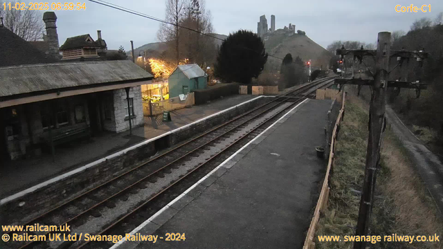 A railway platform with rails running alongside. In the foreground, a stone building with a thatched roof sits to the left, while a green shed is visible nearby. The platform is bordered by wooden fencing and a pathway leads off to the right. Above the platform, an electric pole and wires are present. In the distance, a hill features a ruined castle. The scene is dimly lit, suggesting early morning light.