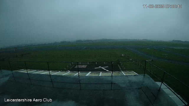 A cloudy and foggy scene from a webcam overlooking a grassy airfield. The view shows a runway in the distance, with a large white cross marked on the ground. A railing can be seen at the forefront, and the image appears dim and overcast, indicating poor visibility. The date and time display in the corner reads November 2, 2025, at 07:00:42.