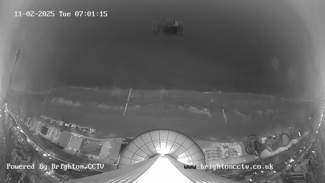 A black and white webcam image capturing a coastal scene. The view includes a sandy beach with gentle waves breaking on the shore. In the distance, an oil rig or similar structure is visible on the water. The foreground shows the outline of a dome structure, possibly a building, with some lights along a promenade area. The top corner displays the time, date, and webcam details. The overall atmosphere is calm and serene.