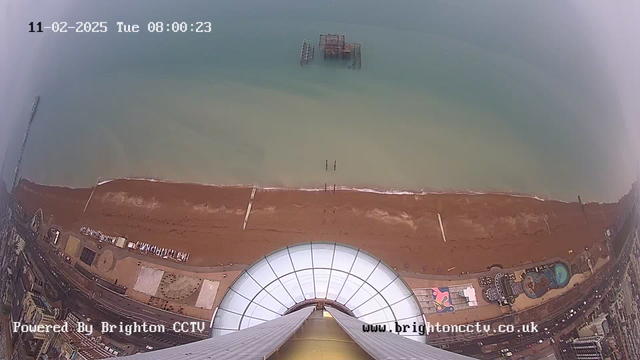 An elevated view of a sandy beach meeting the sea, captured from a webcam. The image shows the coastline with people and structures along the shore. To the left, a pier extends out into the water. Several wooden poles are visible in the water where the pier ends. The beach area features a mix of colors and textures, with some beach amenities and pathways. The sky is light blue, indicating a clear day. A circular glass structure is visible in the foreground, suggesting an observation area or platform. The timestamp at the top indicates the date and time of the image.