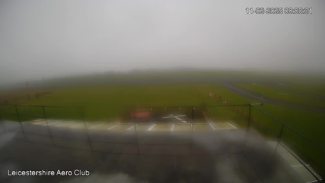 A foggy scene at the Leicestershire Aero Club showing a grassy airfield. The visibility is low, with only partial outlines of the runway and markings visible. A barrier or fence is present in the foreground.