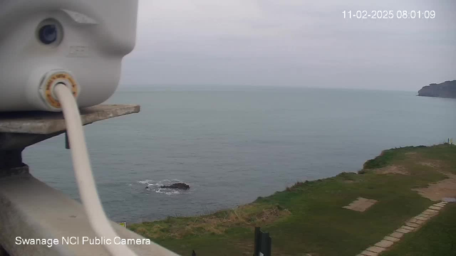 A view of the sea from a coastal location. In the foreground, a white cylindrical object with a cable is visible on a ledge. Below, the water appears calm and slightly rippled, with a rocky outcrop partially submerged. The shoreline features grassy areas and a path made of stone blocks leading away from the edge. The sky is overcast, and the overall scene is serene. A date and time are displayed in the upper right corner.