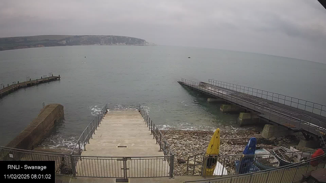 A cloudy sky looms over a calm sea, with visible waves lapping at a concrete ramp leading down to the water. The ramp descends from a flat area bordered by a railing. To the left, a curved jetty extends into the sea, with several boats visible on the surface. In the foreground, there are several colorful kayaks (yellow and blue) stored on the shore next to a rocky area. The scene is tranquil, depicting a coastal environment.