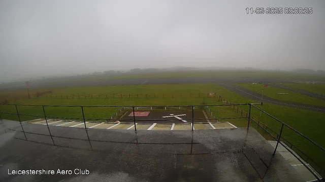 A foggy scene from a webcam at Leicestershire Aero Club. The view shows a grassy area with a parking ground marked in red beneath a railing. In the distance, there are low visibility patches of grass and a hint of runway markings. The sky is overcast and gray, contributing to the lack of clarity in the image.