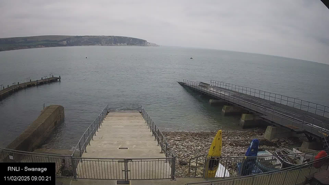 A view of a calm sea under a cloudy sky. In the foreground, there is a staircase leading down to the water from a concrete platform. To the right, a long wooden pier juts out over the water, with railings along its sides. Several boats are docked at the edge, including a yellow kayak and a blue kayak, resting on a rocky shore. The distant shoreline features grassy cliffs.