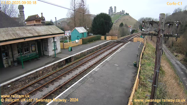 A railway station with a stone building featuring a green bench in the foreground. The platform has two railway tracks running parallel. On the left, there's a wooden fence and a small green building. In the background, a hill rises with ruins at the top, surrounded by trees. The sky is light and overcast. A wooden telephone pole with multiple wires stands on the right side of the image.