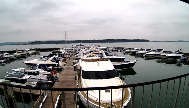 A cloudy view of a marina filled with various boats and yachts docked at the wooden pier. The water is calm, reflecting the boats and the overcast sky. In the background, a distant shoreline is visible, indicating land beyond the marina. The scene conveys a tranquil waterfront atmosphere.