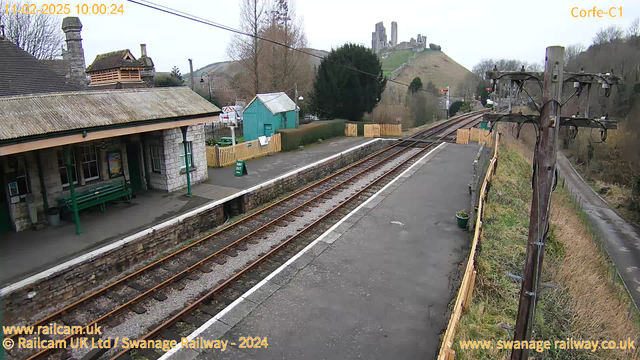 A view of a railway station platform with a stone building on the left featuring a sloped roof and large windows. A long green bench is under the roof awning. The railway tracks are visible in the foreground, leading off towards the right. In the background, a hill with ruins can be seen, partially obscured by trees. A green shed is situated on the platform with a wooden fence running along one side. The weather appears overcast and the scene is empty, with no people present.