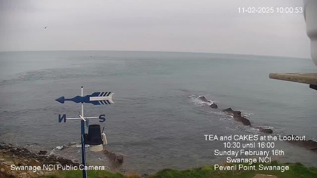 A coastal view from a webcam showing a wide expanse of calm water under a cloudy sky. In the foreground, there is a white and blue weather vane with directional arrows pointing north and south. To the right, rocky outcrops are visible in the water. The bottom of the image includes text promoting a tea and cakes event at a lookout point in Swanage, along with the date and location details.