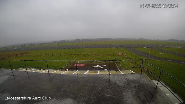 A view from a webcam shows an expansive, grassy field under a cloudy sky. In the foreground, there is a railing, and below it is a tarmac area with a white cross marking on the ground. The background features a runway and a distant horizon where trees are faintly visible. The scene appears dreary and overcast, indicating possibly rainy weather. The image is timestamped, showing the date and time in the corner.