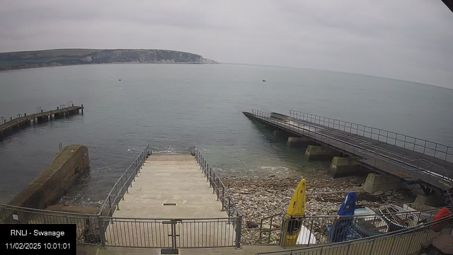 A cloudy daytime view of a coastal area featuring a calm sea. In the foreground, there is a concrete ramp leading down to the water, bordered by a metal railing. To the right, a wooden pier extends over the water. On the shore, there are several kayaks in various colors, including yellow and blue, resting on rocky ground. The background shows cliffs and hills in the distance, under a gray sky.