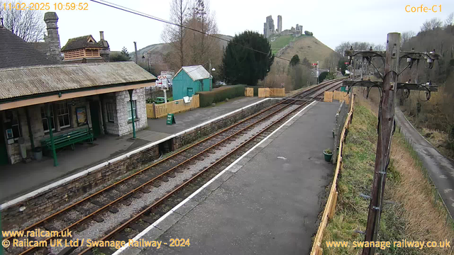 A view of Corfe Castle station with a platform in the foreground featuring a stone building and a roof with a slight slope. There is a green wooden bench against the wall and a green shed to the left. A sign labeled "WAY OUT" is visible on the platform. The railway tracks run straight through the middle of the scene, bordered by gravel and a low stone wall. In the background, the hill rises with the ruins of Corfe Castle perched on top, surrounded by bare trees and shrubbery. The sky is overcast and gray.