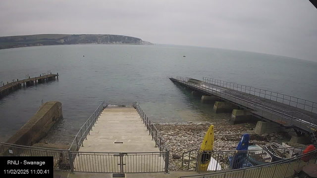 A view of a calm sea with a rocky shoreline. In the foreground, there are concrete steps leading down to the water, bordered by a metal railing. To the left, a wooden pier extends into the water. In the center, there are various colorful kayaks stored upright, with a yellow one prominently displayed. In the distance, a rocky cliff rises along the coast under a gray, overcast sky. The scene conveys a tranquil atmosphere.