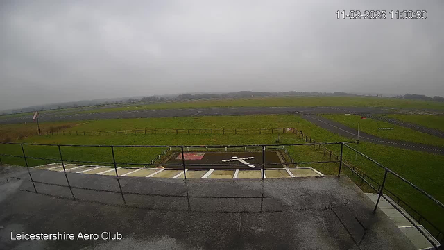 A cloudy, overcast sky overlooks a grassy airfield. In the foreground, there's a railing with a wet surface, likely from recent rain. The airfield stretches out, showcasing a large expanse of green grass intermixed with a dark runway and a marked landing area in the center. A windsock flutters on the left side, indicating wind direction, while a small red flag stands nearby. The date and time display is located in the upper right corner of the image.